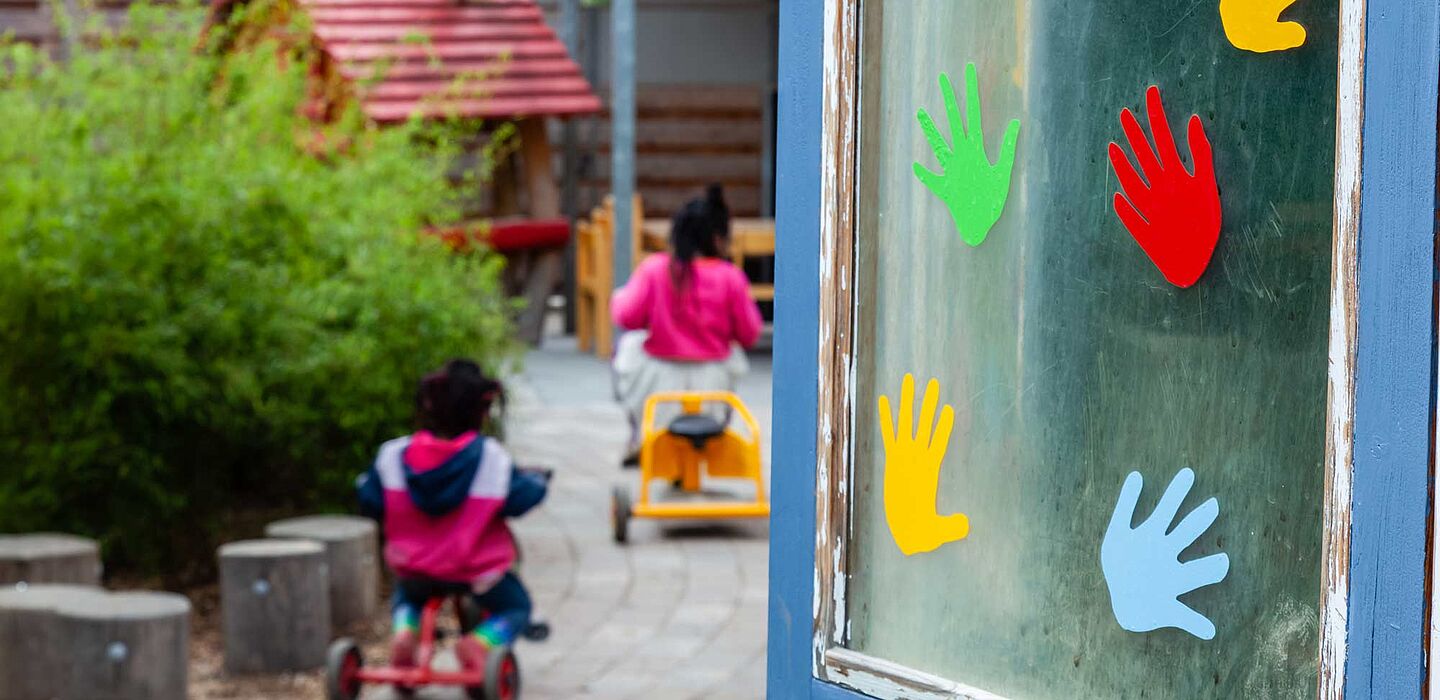 Aussenaufnahme, links laufen Kinder in Richtung Kita, rechts sind an einer Fensterscheibe bunt Handabdrücke sichtbar