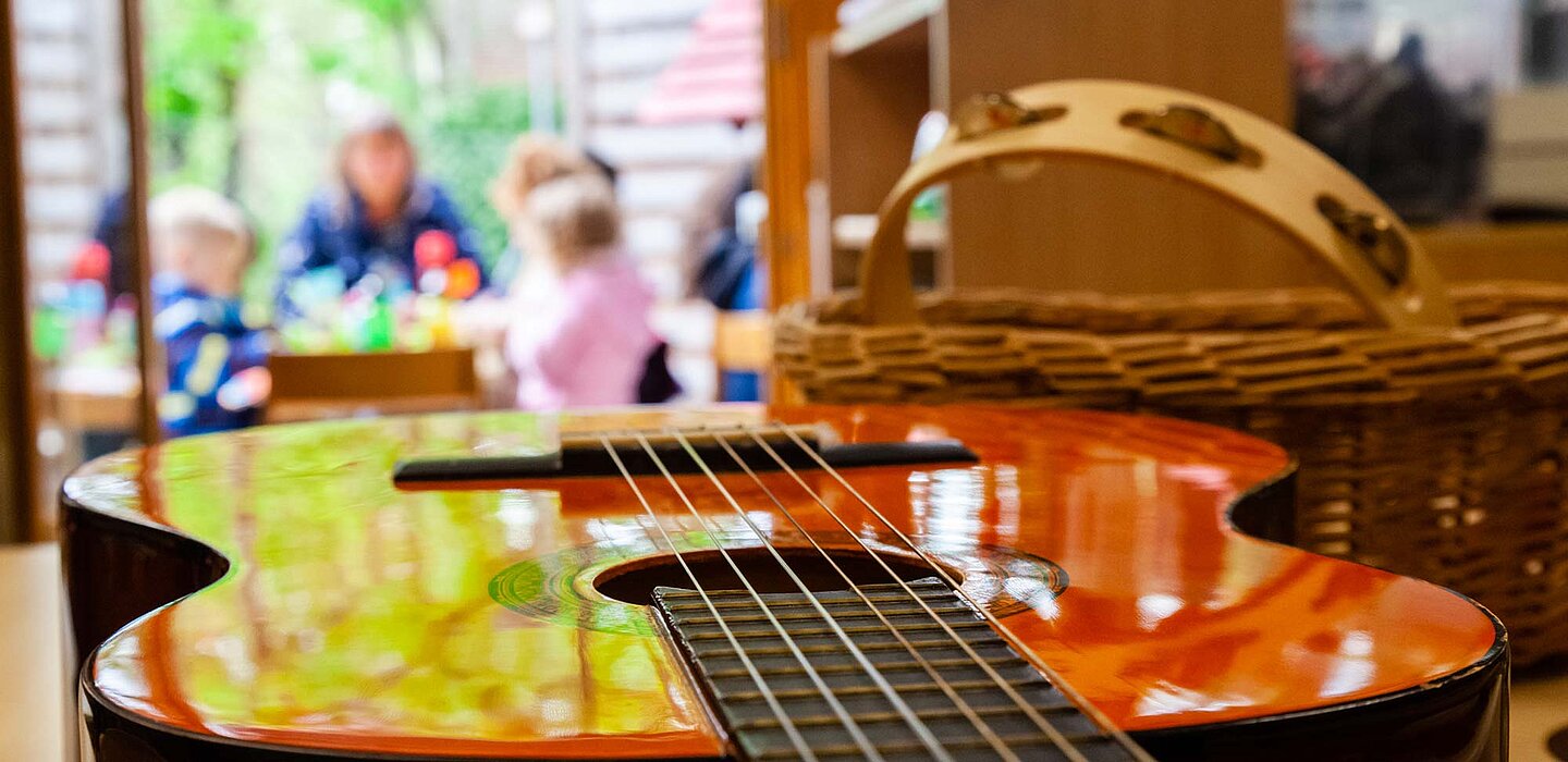 Im Vordergrund eine Gitarre, im Hintergrund eine Gruppe Kinder mit einer Erzieherin