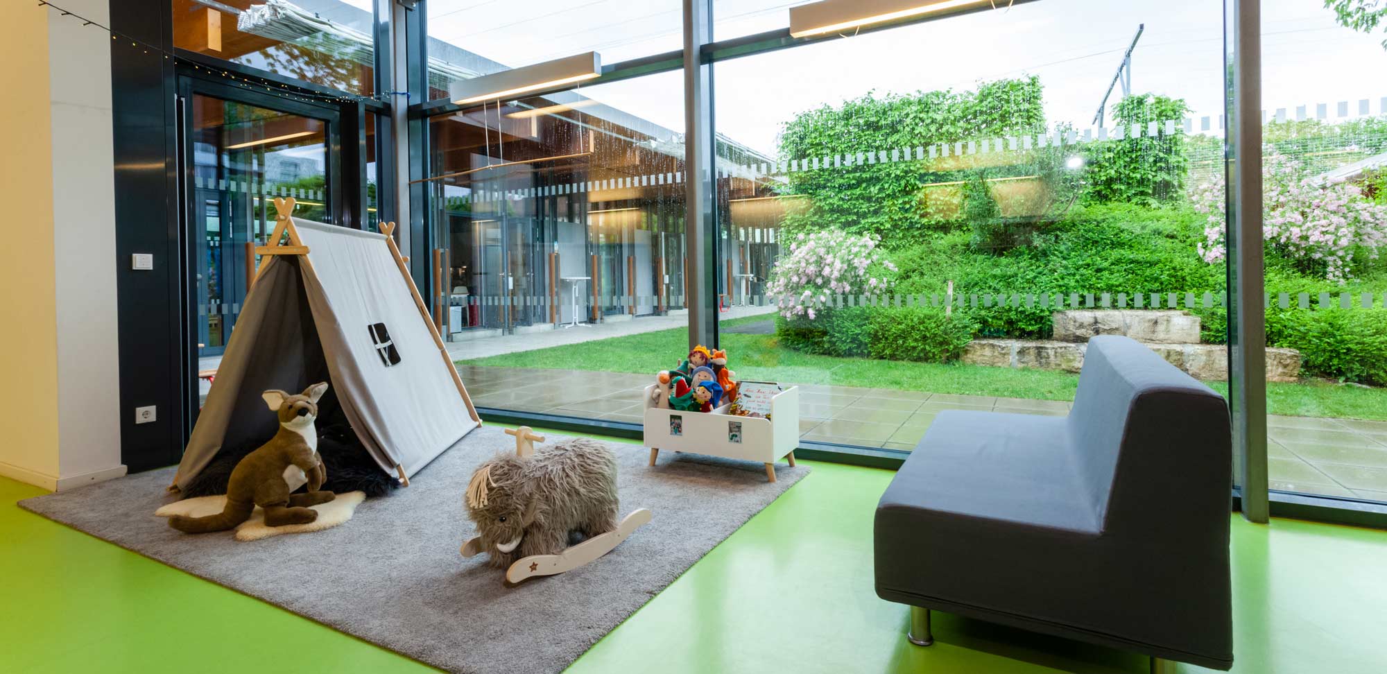Sicht auf eine Spielsituation in der Betriebskita Sailerstraße. Links ein Zelt auf einem Teppich und rechts eine Couch. Im Hintergrund eine große Fensterfläche die den Innengarten zeigt. 